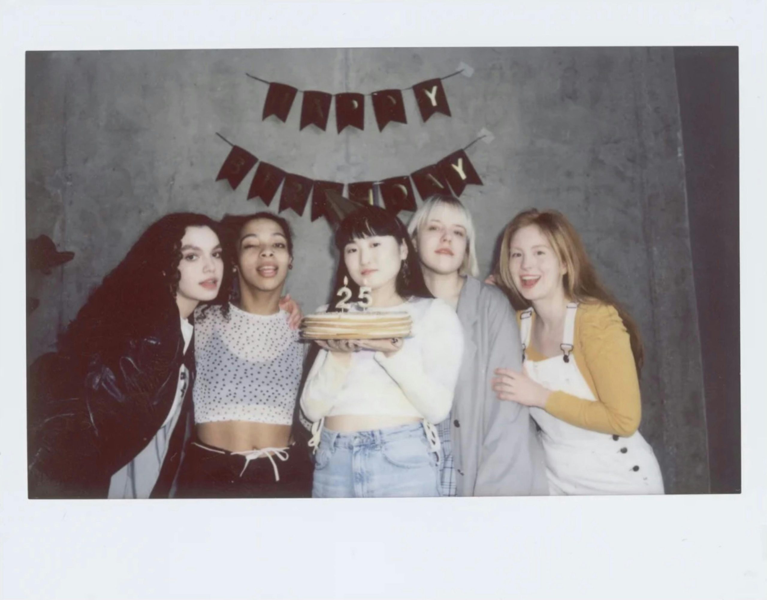 Five friends celebrating a 25th birthday indoors with cake and decorations.